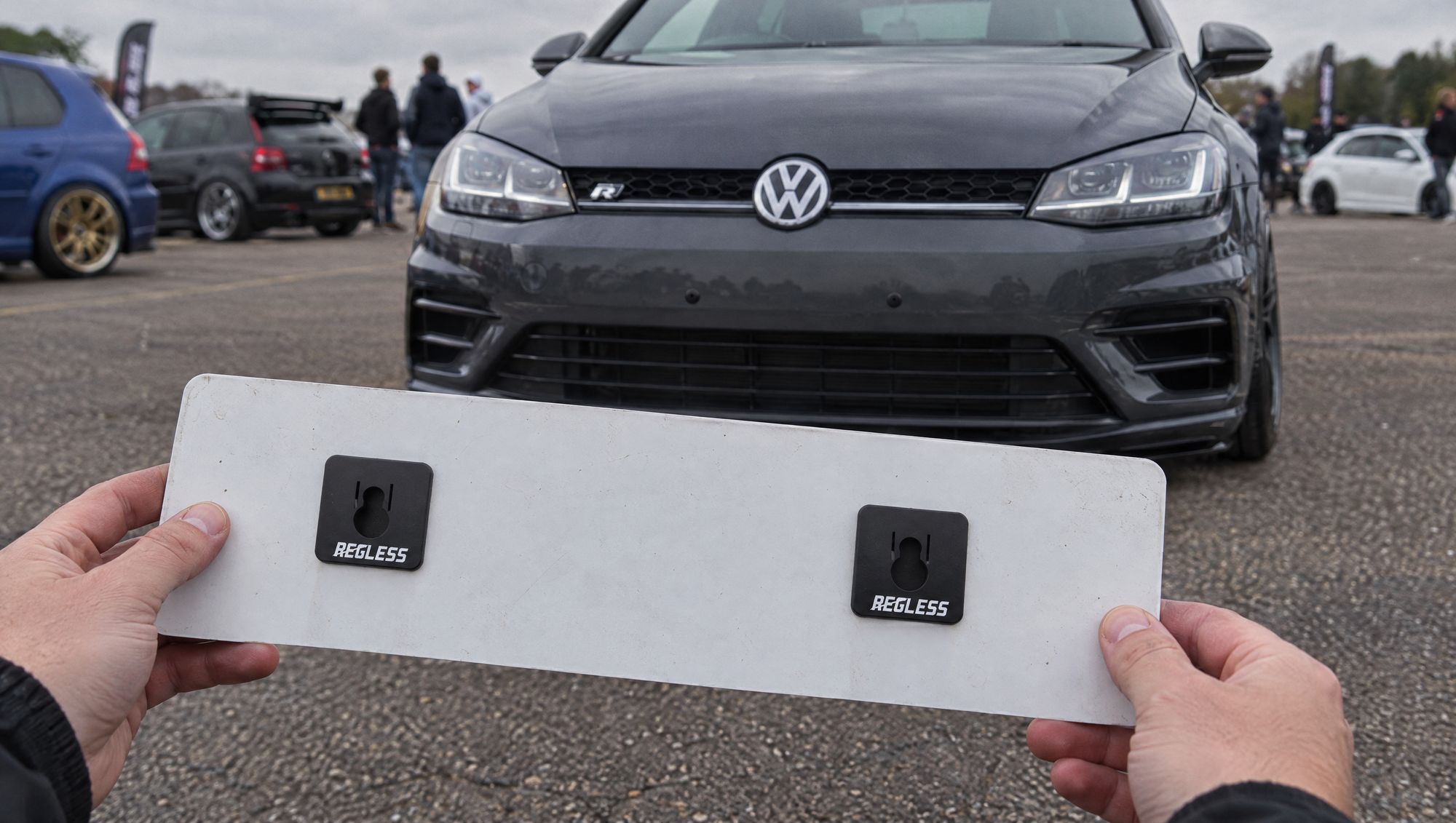 Owner at a meet holding the front plate turned round, two Regless squares on its back; the plateless bumper behind shows just two small pegs